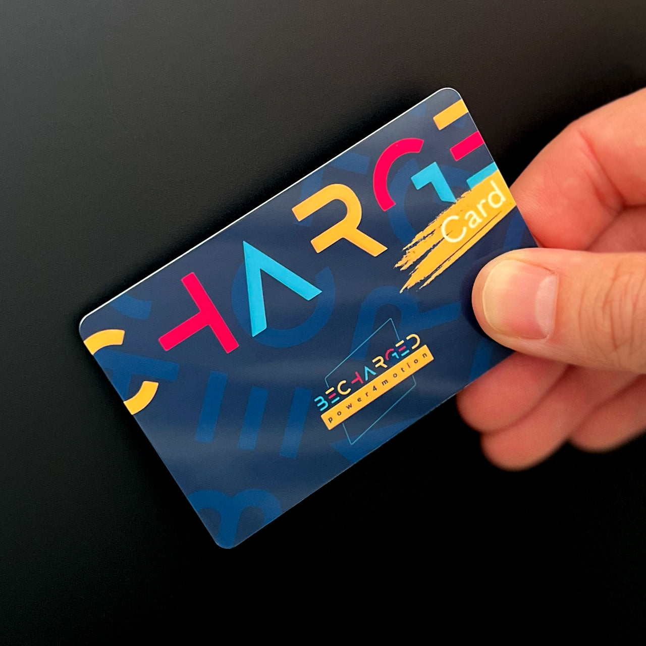 Eine Hand hält die becharged RFID-Karte an eine Oberfläche. Die RFID-Karte ist dunkelblau, darauf ist in der oberen Hälfte von links nach rechts in großen gelben, roten und hellblauen Buchstaben "Charge" geschrieben. Unter den letzten beiden Buchstaben ist ein gelber Pinselstrich auf dem in weißen Buchstaben "Card" geschrieben steht. In der unteren Hälfte ist das bunte Logo "becharged power4motion" aufgedruckt.