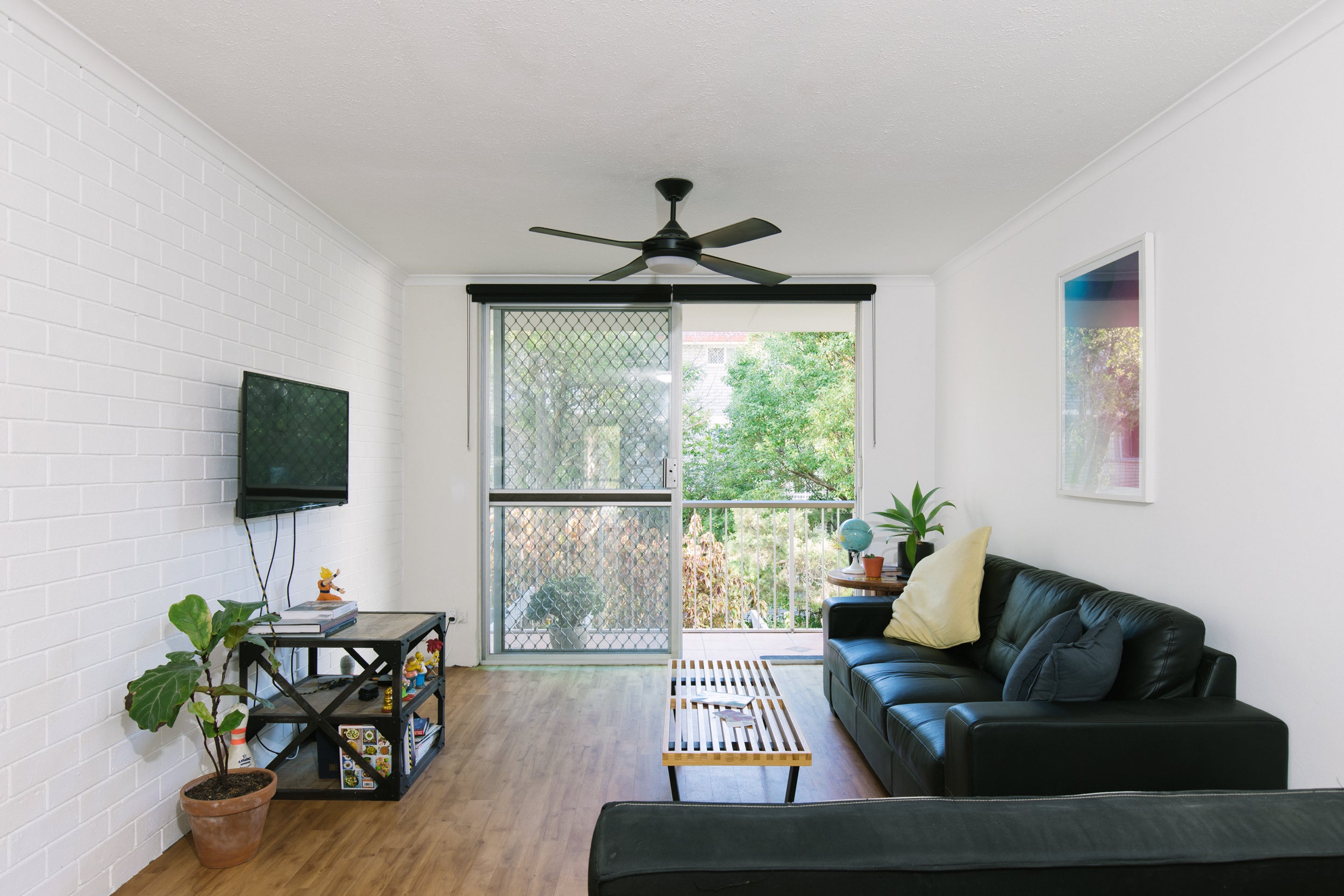Ein weißer Raum mit einem großen bodentiefen Fenster auf der gegenüberliegenden Seite. Rechts an der Wand steht eine schwarze Couch, davor ein Couchtisch. An der linken Wand hängt ein Fernseher über einem Regal mit drei Etagen. An der Decke hängt ein Ventilator.